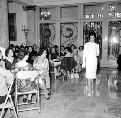 Eva Sámano de López Mateos y demás mujeres observando a modelo desfilar durante exhibición de modas en beneficio de la biblioteca de la Presidencia, en la cd. de México