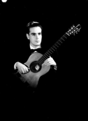 Manuel López, sentado con una guitarra apoyada en sus piernas, viste de smoking, retrato de frente