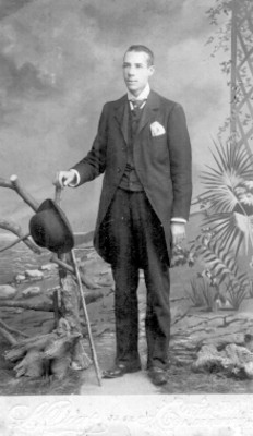 Hombre posa de pie con la mano derecha sobre un tronco, retrato