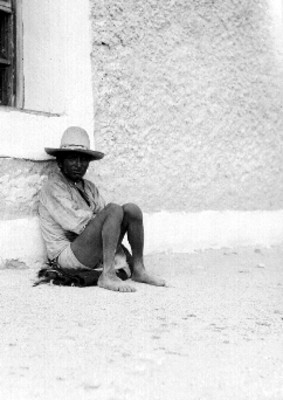 Tarahumara con sombrero sentado junto a la pared de una casa, retrato