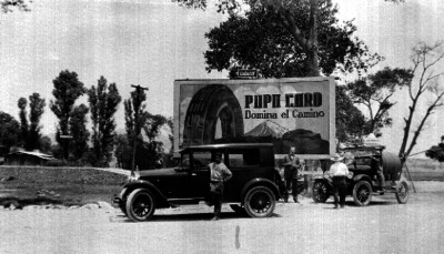 Hombres y automóviles en una carretera junto a un letrero de la Popo Card