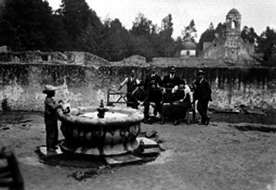 Hombres y mujeres vestidos elegantemente sentados en una banca junto a una fuente durante un día de campo en El Desierto de los Leones, retrato