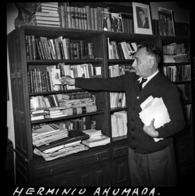 Herminio Ahumada de pie junto a un librero observa fotografía
