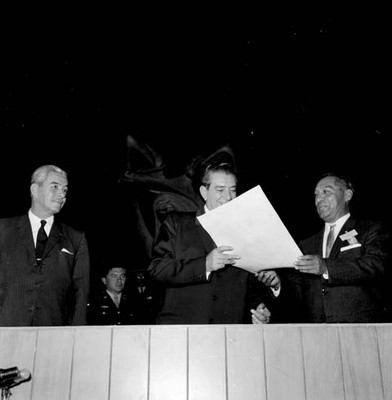 Adolfo López Mateos recibiendo un documento de parte de individuo y los observa Salomón Gónzalez Blanco
