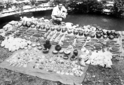 Hombre frente a lote de objetos y restos humanos hallados en el cenote sagrado de Chichén Itzá, reprografía