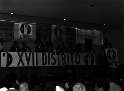 Raúl Noriega y politicos en presídium, ante las palabras de un hombre, durante una asamblea del PRI, en un auditorio