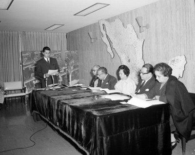 Eva Samano de López Mateos junto a hombres y mujer revisan documentos en un salón durante conferencia