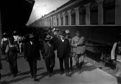 Alvaro Obregón acompañado por funcionarios y militar, caminando en andenes de una estación del ferrocarril