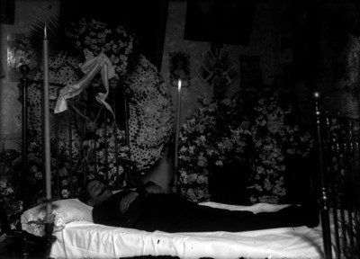 Cadáver de un hombre sobre una cama, rodeado de flores