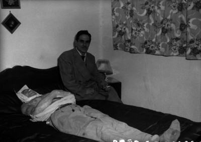 Hombre vestido de traje sentado a la orilla de una cama junto a una figura humana de cera